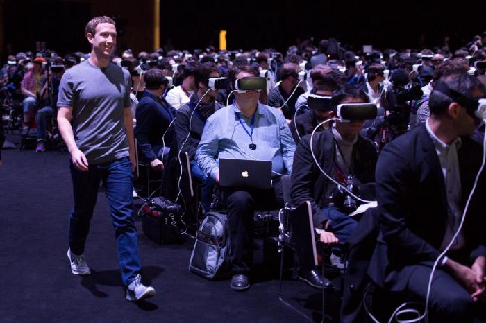 Mark Zuckerberg walks, smiling, through a sea of seated men wearing VR headsets.