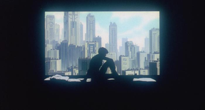 Motoko Kusanagi sits up in bed in a dark room, framed in silhouette against the large window behind her. Through the window, the towers of a brightly sunlit city can be seen.
